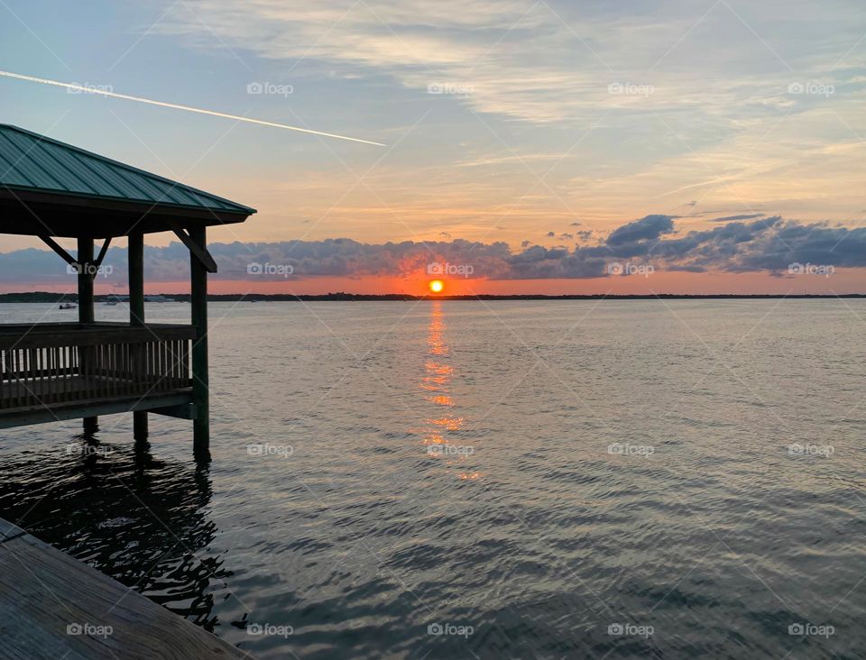 Sunset In The Horizon With Deep Orange Color Sighting By The Gazebo On The Dock By The Island Reflecting On The Water.