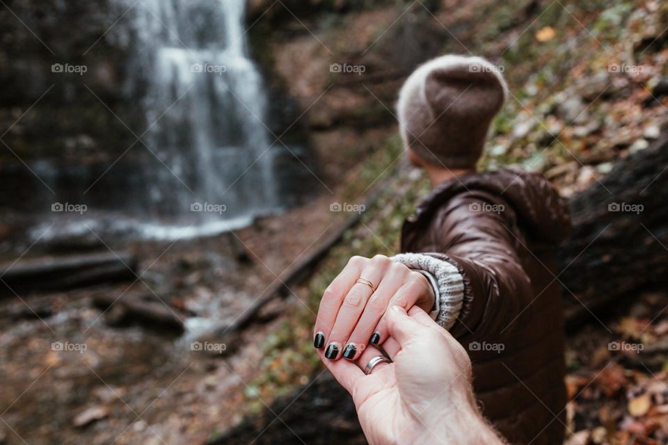 A couple walks hand in hand towards a waterfall in the forest.