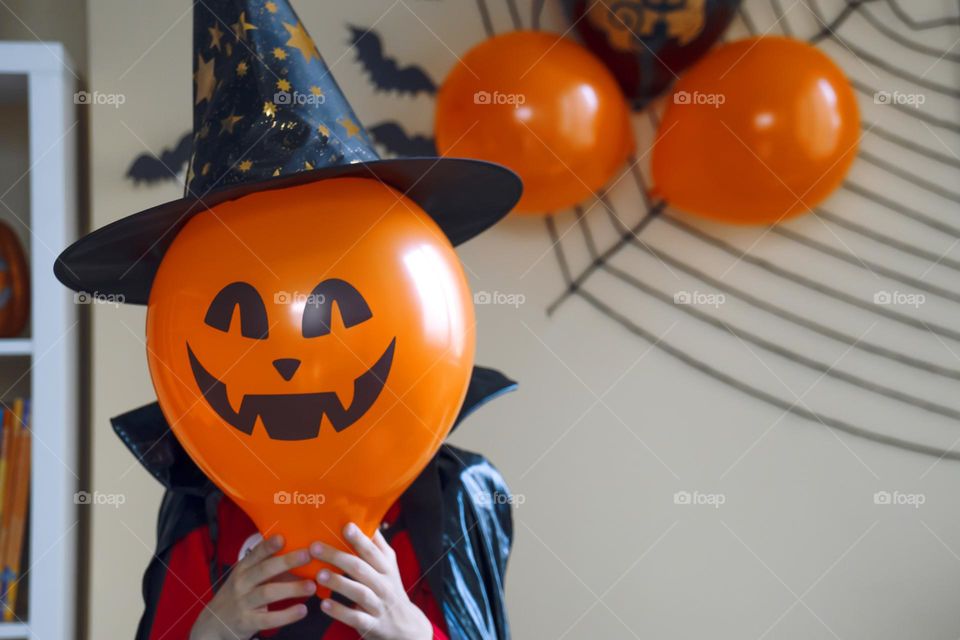 Boy in a black cape and a witch's hat holds a balloon with the image of a pumpkin jack in his face