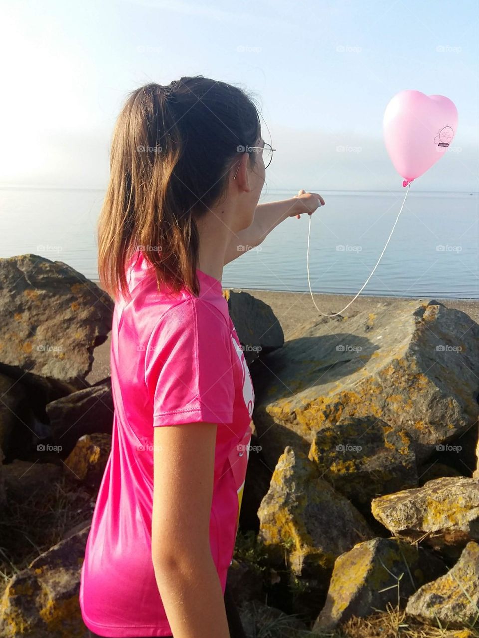 From the back, a girl wearing a ponytail and wearing a bright pink t-shirt holds a pink balloon in the shape of a heart.