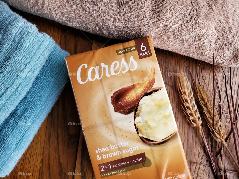 The new look Caress bar soap shea butter and brown sugar six pack, next to a tan towel and light blue towel on a wood surface whith a natural accent on the right.