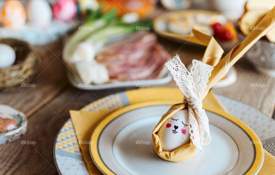 Easter egg decorated as a bunny on plate with delicious traditional easter foods on table