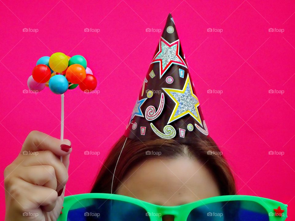 Party time!!  
Girl in party hat and oversized sunglasses holding a mini bouquet of balloons against a bright pink background. 