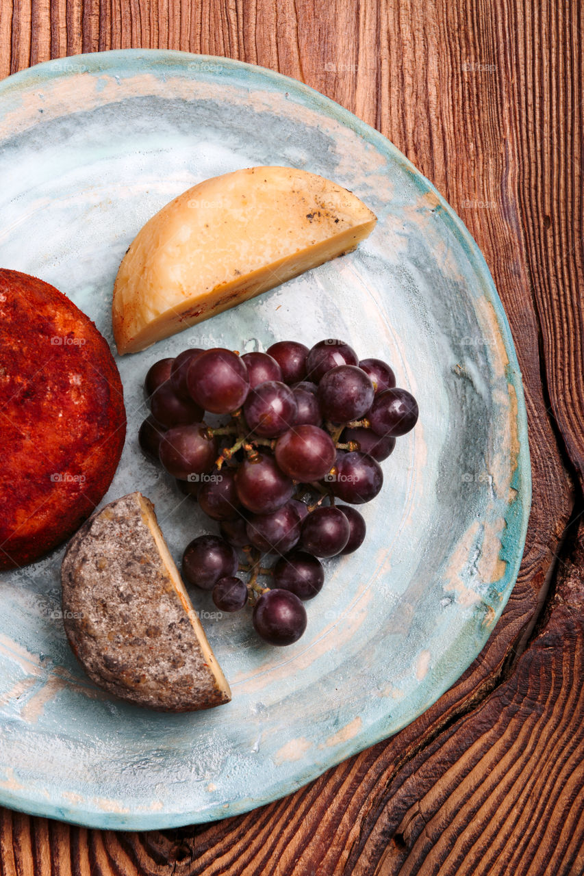Cheese and black grapes on handmade blue pottery plate on old wooden table from above