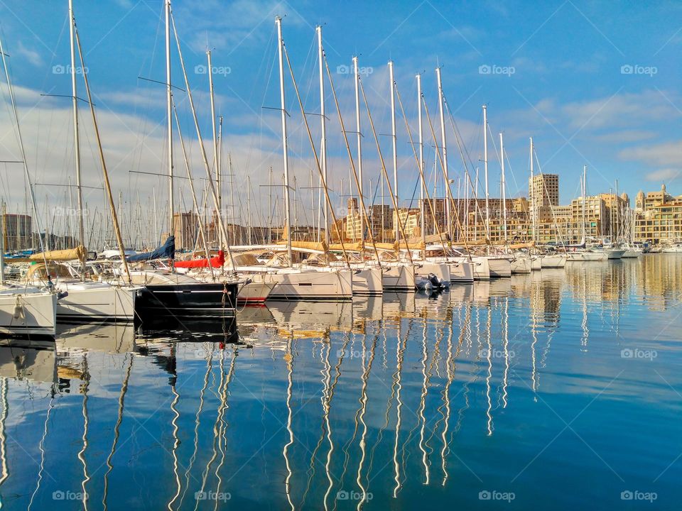 Old port of Marseille, France. In the background the buildings of the city, a blue sky with clouds and more closely boats with their silhouette reflected in the water like a fishbone.