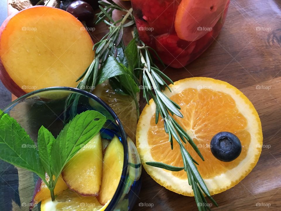 Making healthy flavored waters from garden herbs and summer fruit conceptual wellness and self care healthy drinks background with orange, blueberry, Rosemary., mint, sliced orange and peach on wood tray