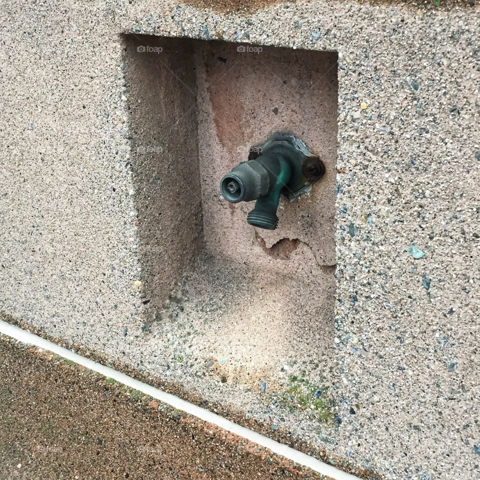 A spigot set in a cement wall. The spigot is made of greenish metal in the cement wall is gray.