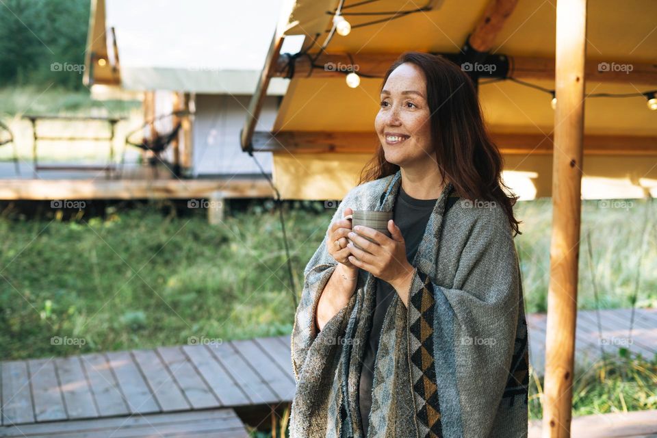 Young brunette woman in poncho drinking tea and relaxing in glamping in nature