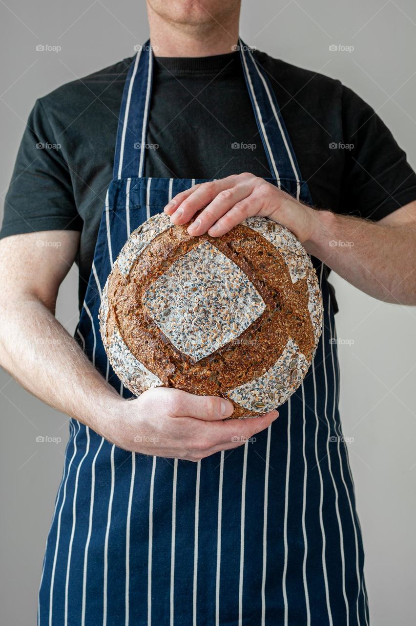 Close-up at bakers hands holding a loaf of sourdough bread in front of him.