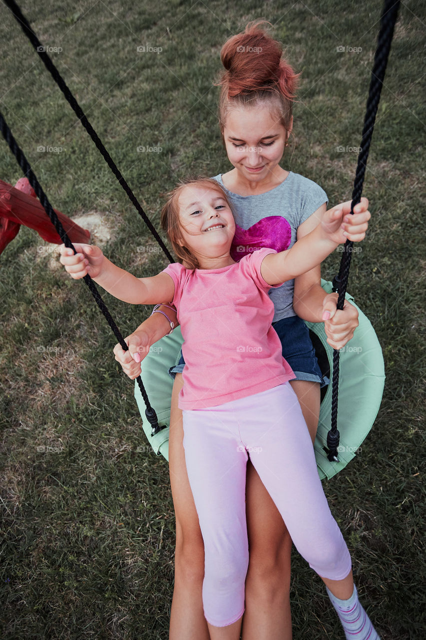 Teenage girl playing with her younger sister in a home playground in a backyard. Happy smiling sisters having fun on a swing together on summer day. Real people, authentic situations