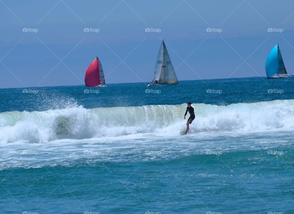 Come Sail Away. Colorful sailboats drift by as a wave closes out on a surfer. Sitting ashore...I dream of sailing off into the sunset.