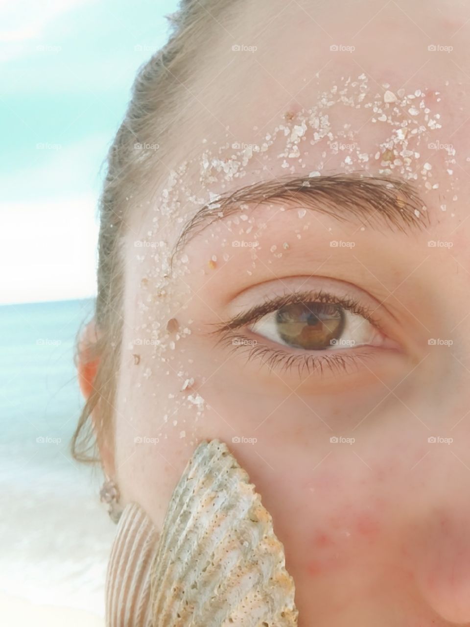 a beautiful girl with a sandy brow and a pattern of seashells along her cheek with ocean behind her