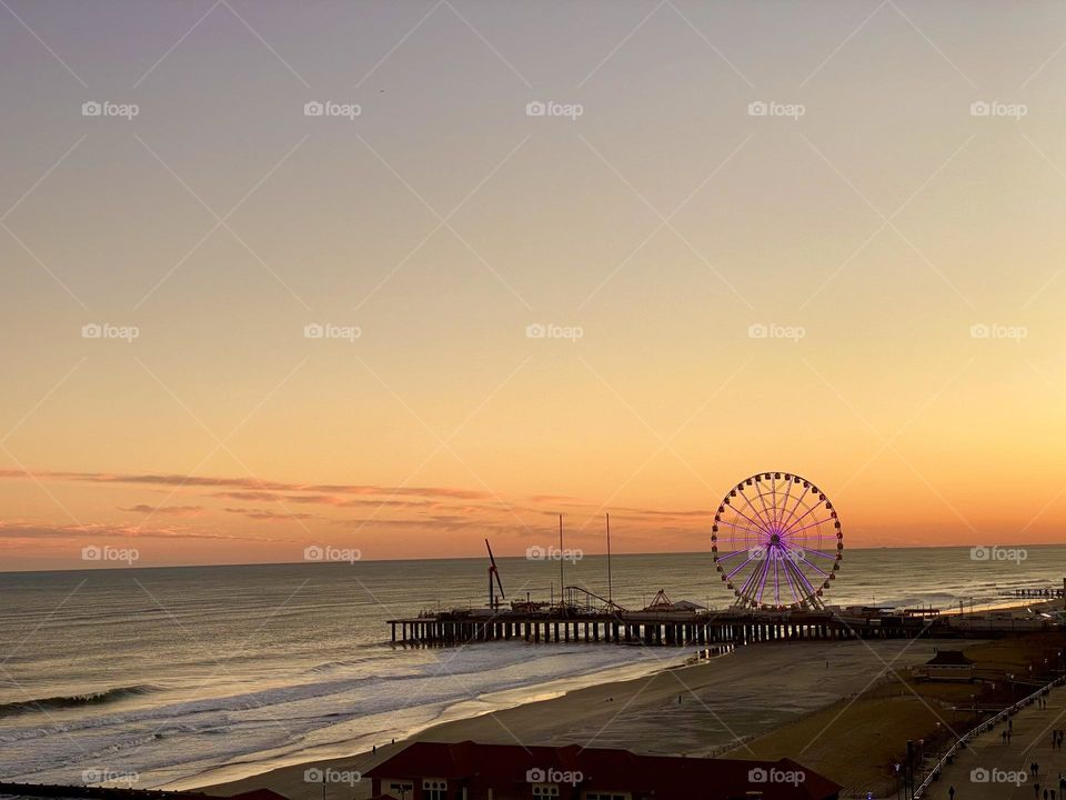 Sunset over the pier down the shore illuminating the Ferris wheel and showcasing the beautiful waves