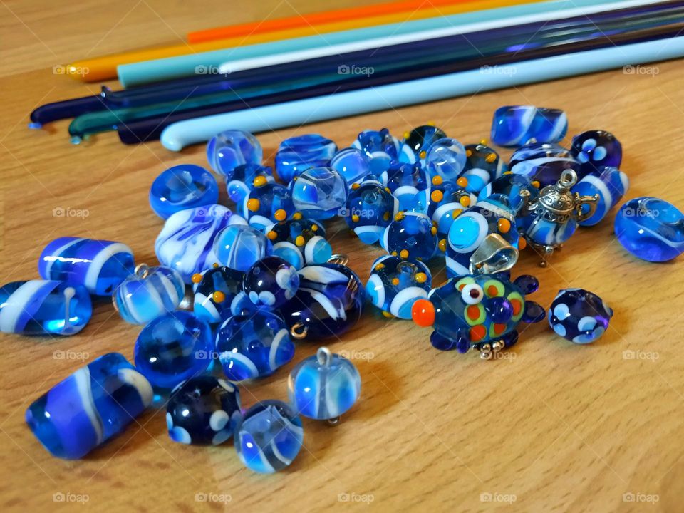 Multicolored handmade glass beads (lampwork). Mainly blue beads, one fish bead. At the background, glass rods used for the handicraft.