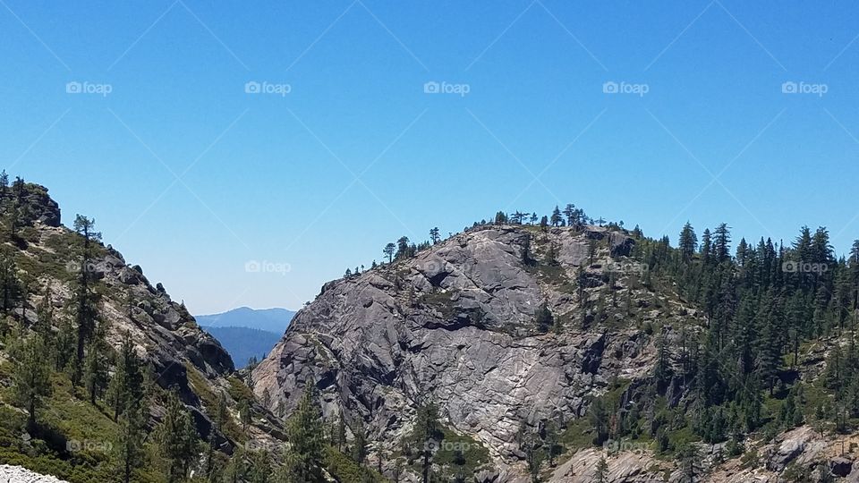 a rocky mountain with trees and a bright blue sky.