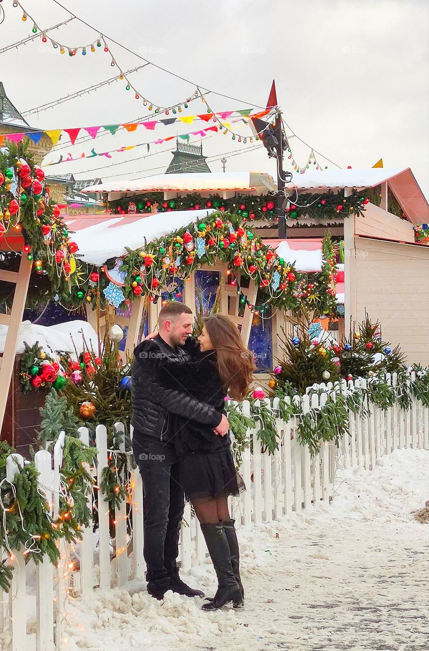 The main motivation of a person.  Love, create a family and have children.  A couple of lovers embrace in a snowy square.  The look of lovers.  Garlanded tents in the background