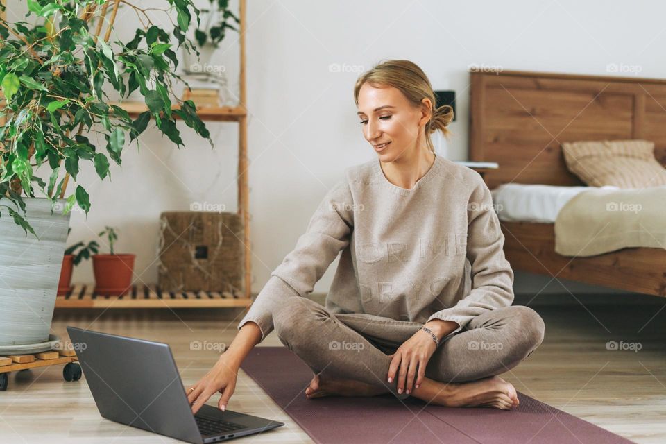 Beautiful blonde young woman in comfortable sport wear practice yoga online in cozy bedroom at home