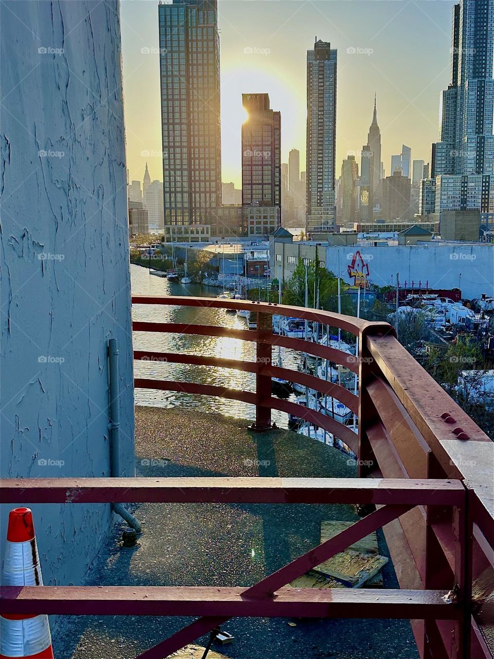 This is the center spot on the „Pulaski Bridge“ at „Newtown Creek“ connecting „Greenpoint“, Bklyn to LIC in the intense setting evening sun. Straight ahead in the distance we see „Manhattan“ incl. the „Empire State Bldg“.  2024. Hypnotic Productions