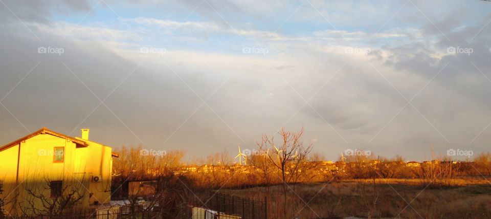 A landscape under the setting sun with a house in orange, blue cloudy sky and windmills on the background