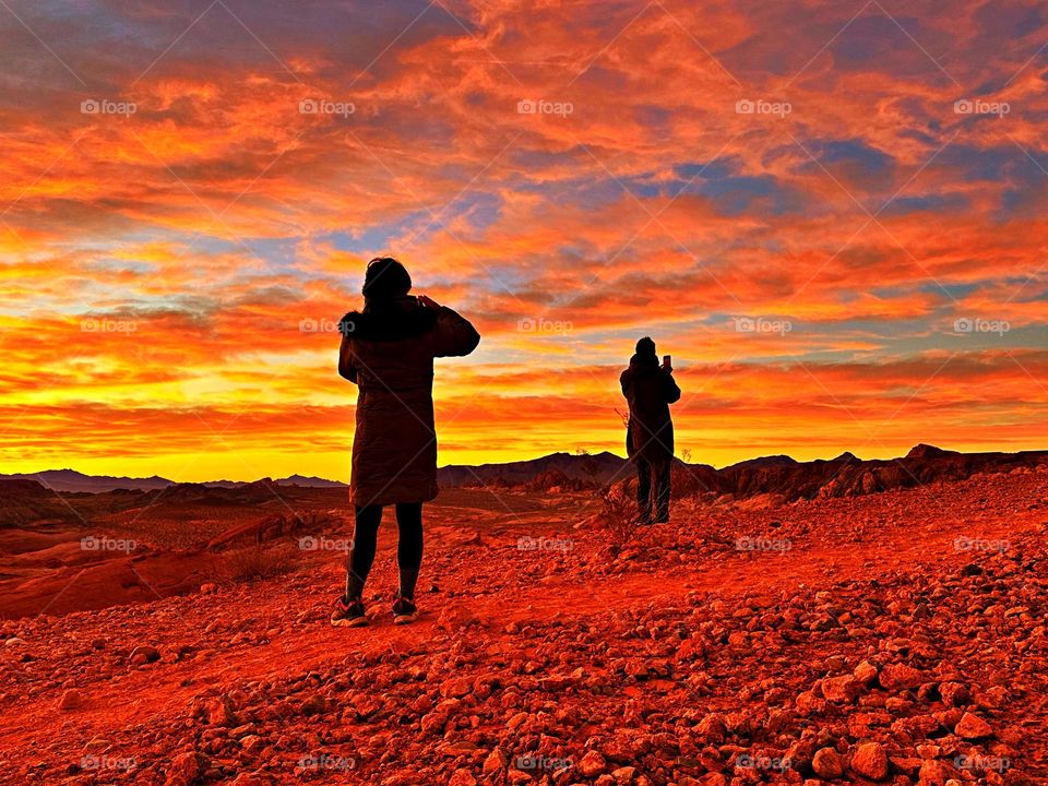 Battle: Summertime vs Spring - Silhouette of two people photographing a vibrant, summer sunrise at Valley of Fire State Park in Nevada.