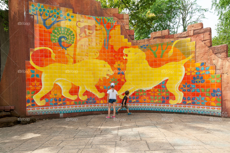 Children admiring colorful tiled wall depicting tale about two fighting male lions.