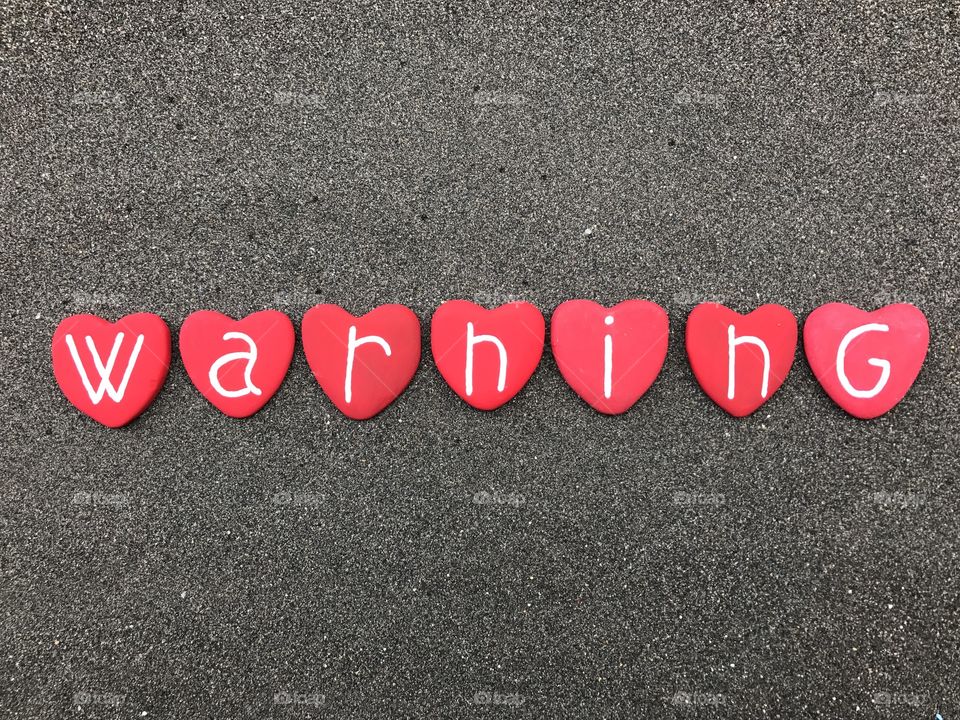 Warning word with red colored heart stones over black volcanic sand