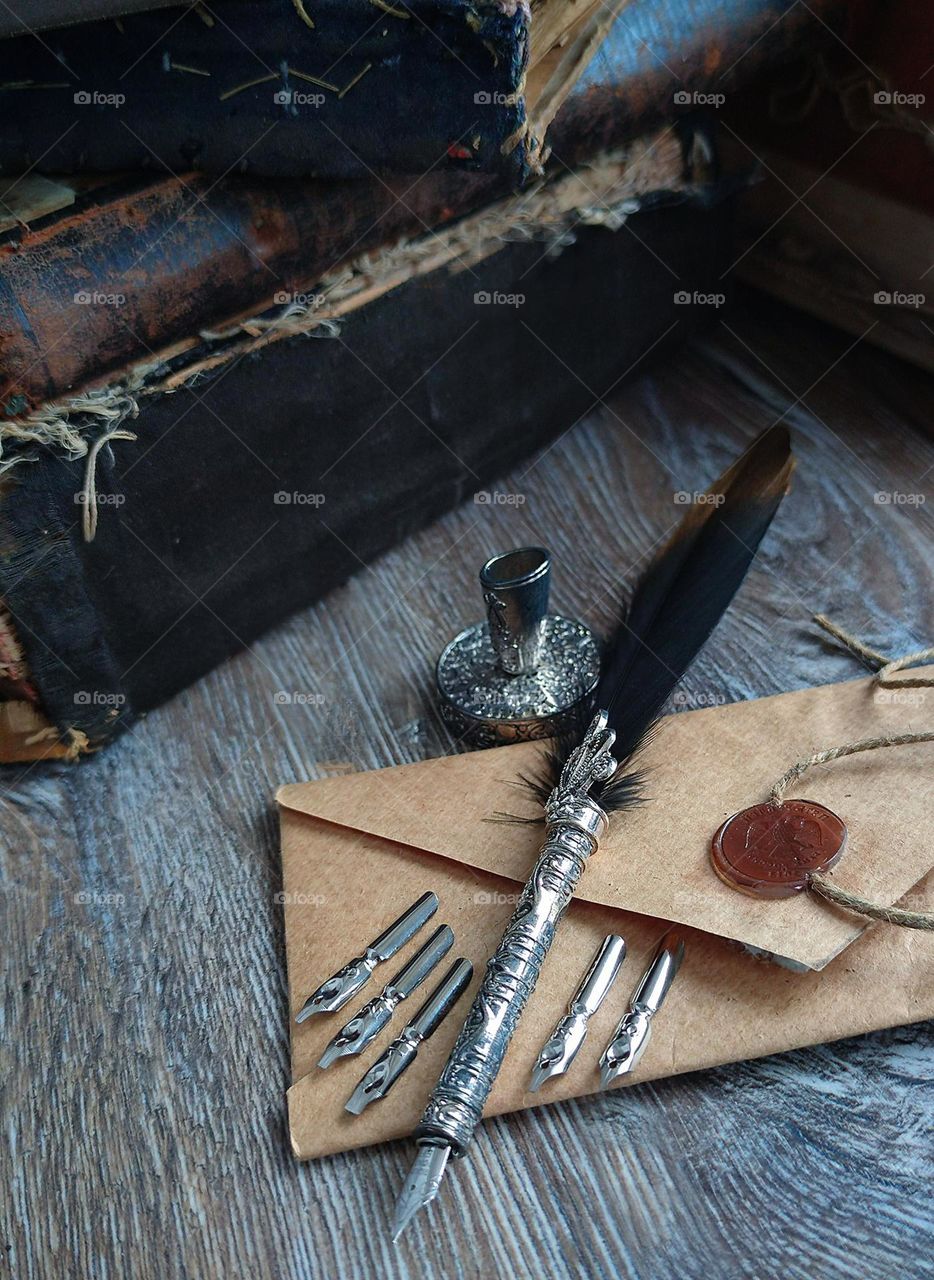 On the wooden one lies a brown envelope with an official wax seal.On the envelope is a metal fountain pen with a bird feather.  Next to the envelope are metal nibs with different tips.Near the envelope is a metal inkwell.Old books in the background