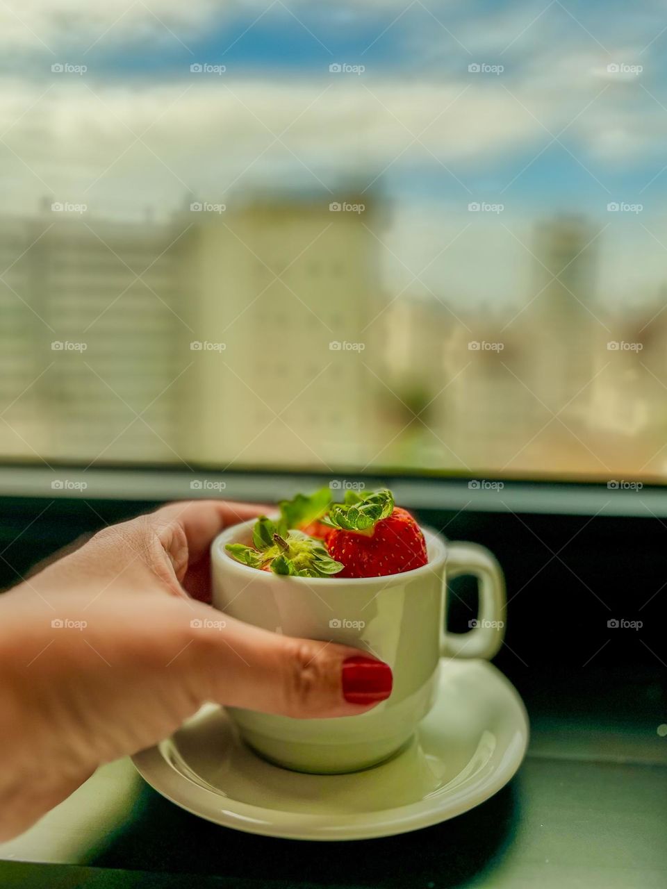 The image shows a female hand holding a small cup with fresh strawberries and some mint leaves, while the blurred background reveals a blue sky with clouds and an urban environment, transmitting a moment of freshness and taste for gastronomy.